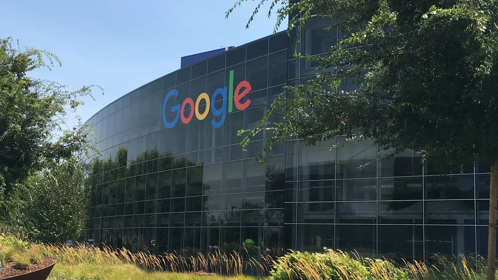 Стеклянное здание штаб-квартиры Google в Маунтин-Вью с разноцветным логотипом за травой и деревьями